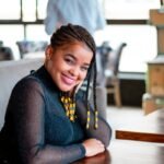 A cheerful woman with braided hair sits in a cozy café, exuding warmth and friendliness.