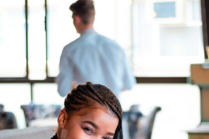 A cheerful woman with braided hair sits in a cozy café, exuding warmth and friendliness.