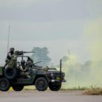 Army soldiers operating a military vehicle during a field exercise with smoke effects.