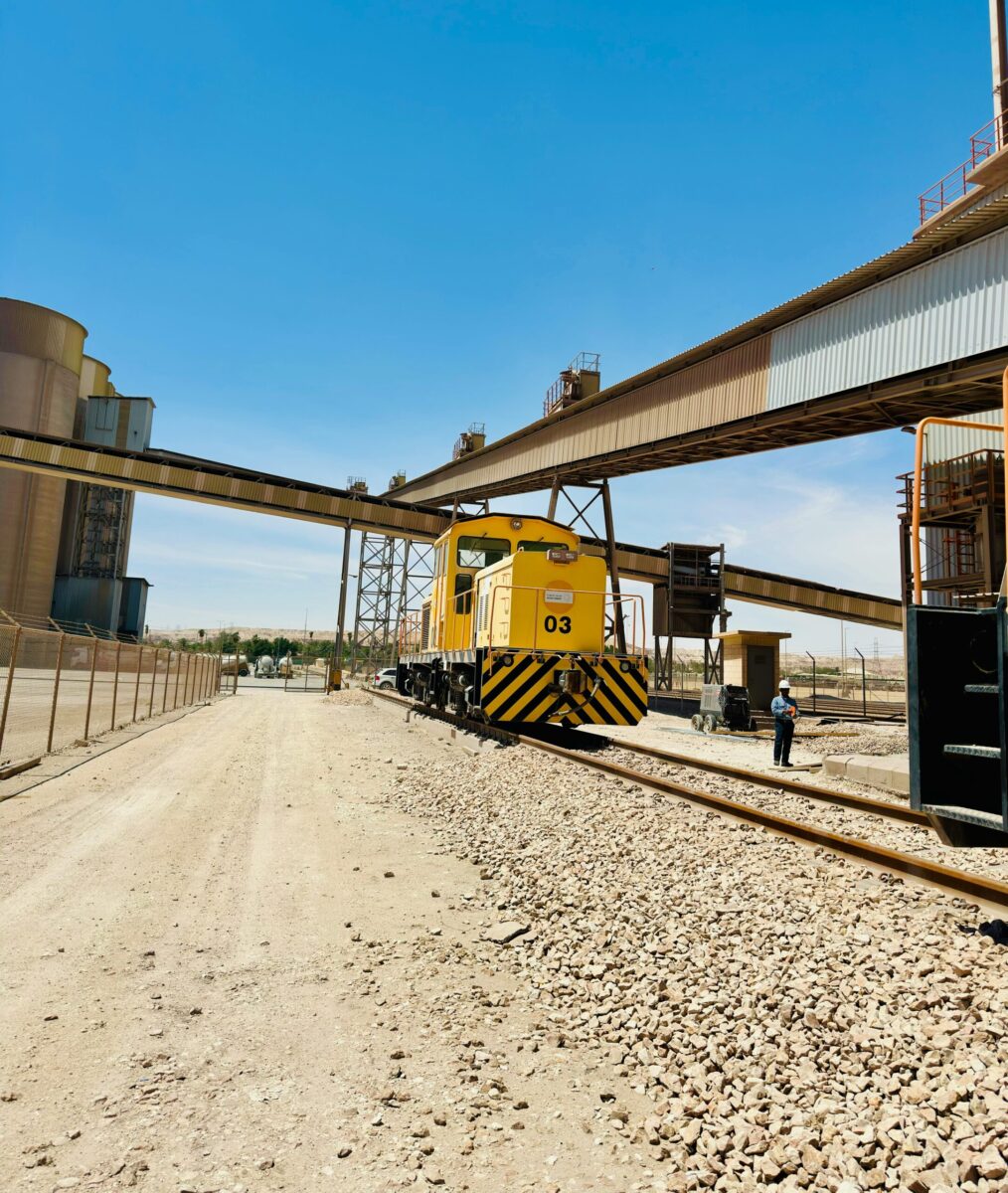 A yellow industrial train at a cement factory track in Saudi Arabia under a clear blue sky.