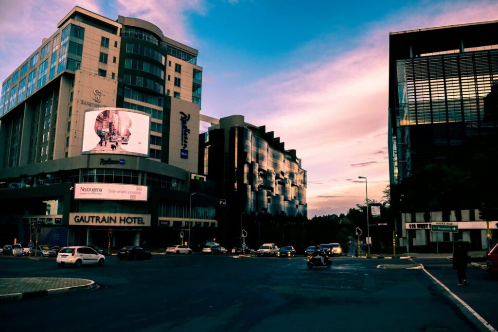 A bustling city street with modern skyscrapers and vehicles at sunset, Gautrain Hotel visible.